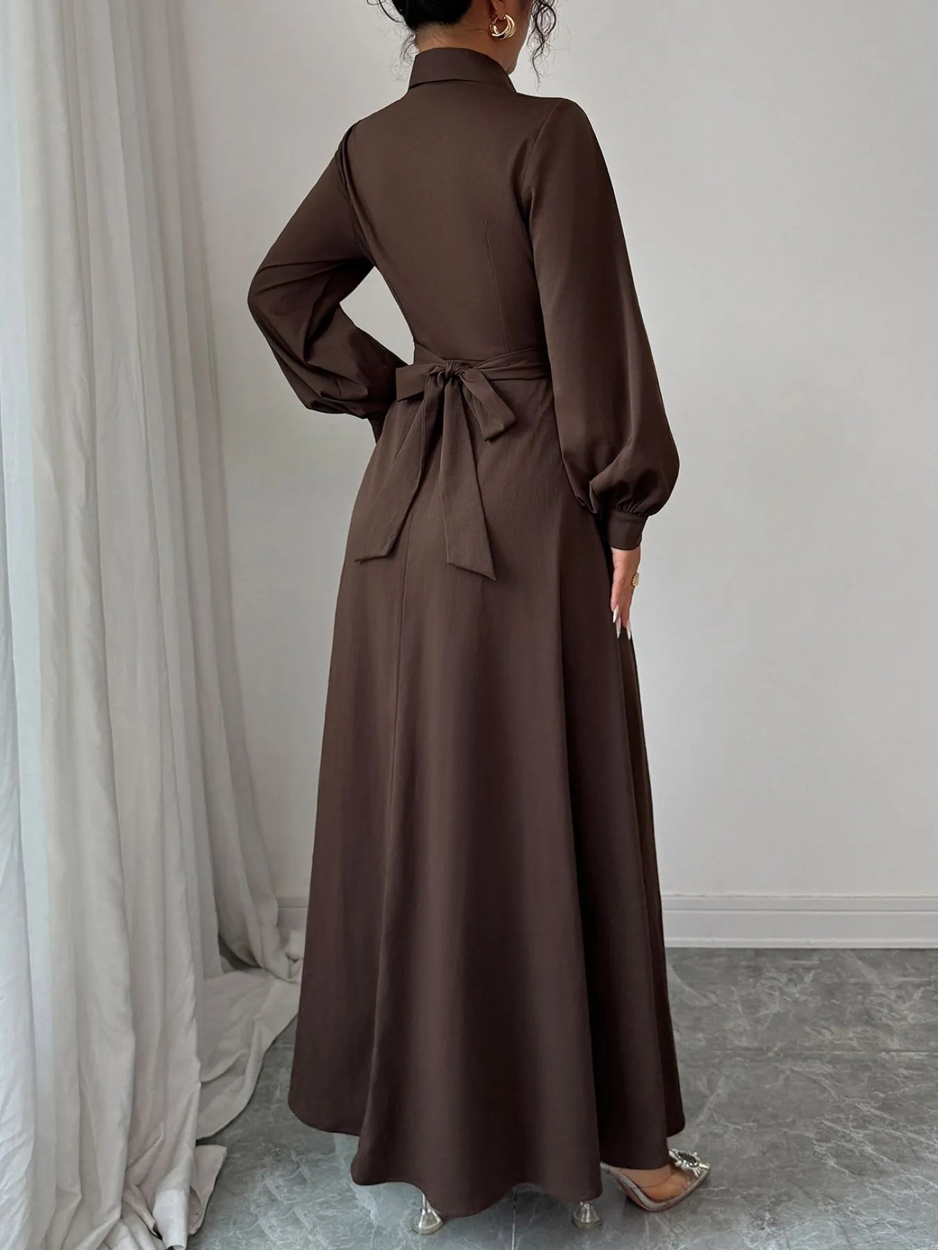 Model wearing a brown long sleeve maxi shirt dress with tied waist bow and puffed sleeves back view