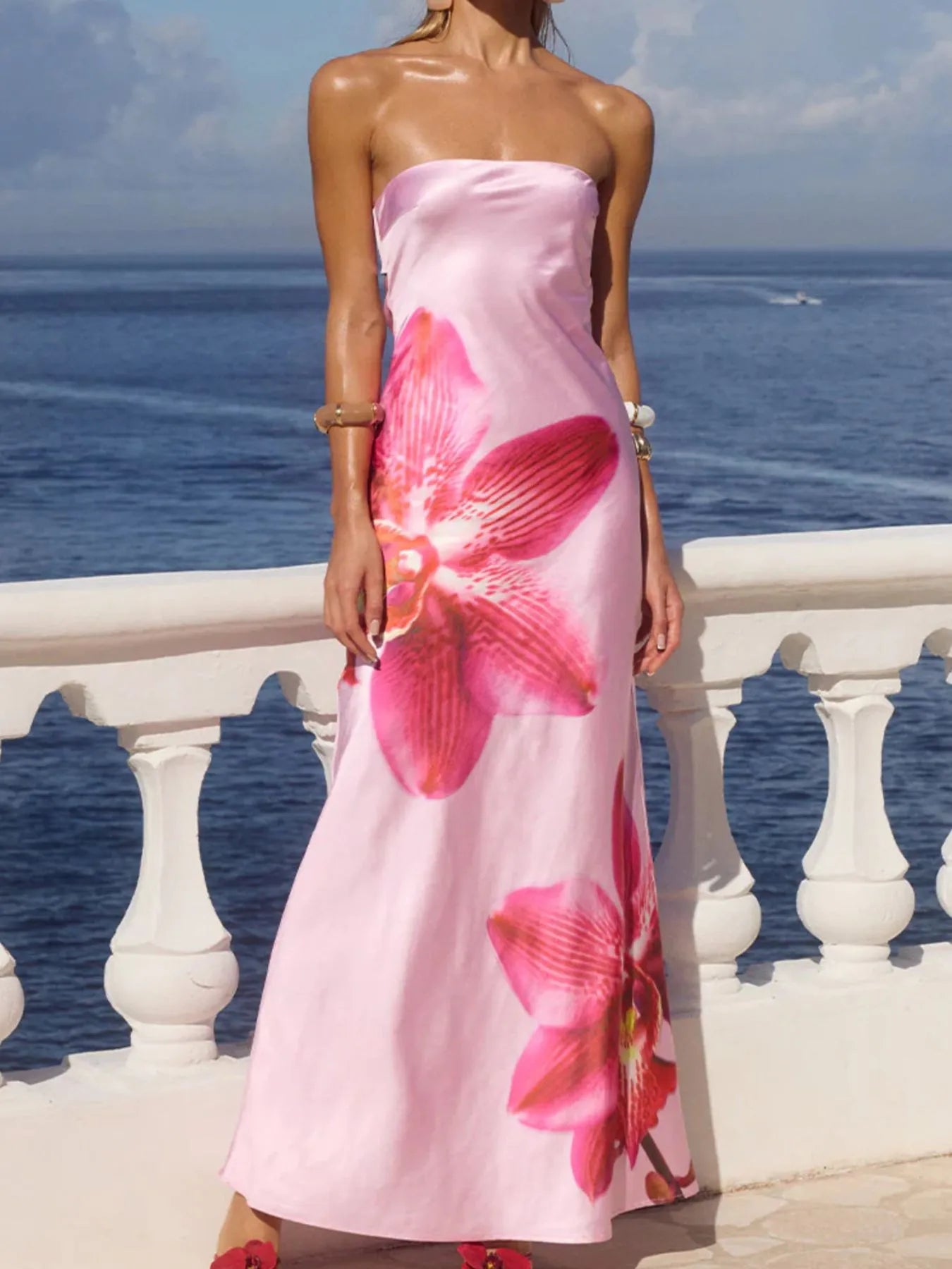 Model wearing printed tube maxi dress strapless pink satin with large orchid floral print on seaside balcony