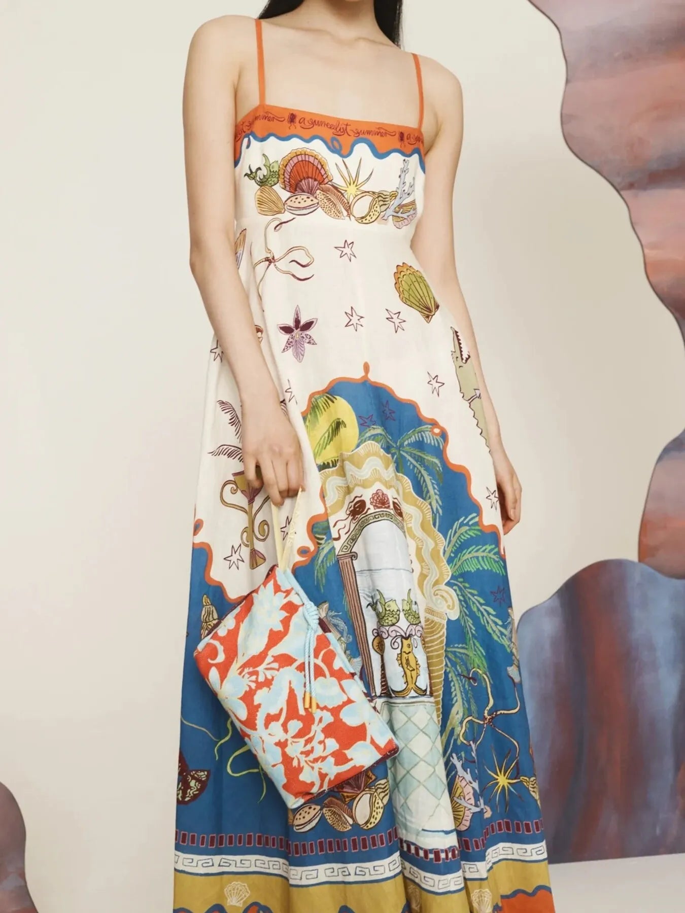 Model wearing a colorful printed square neck maxi dress with thin straps and coral patterned clutch