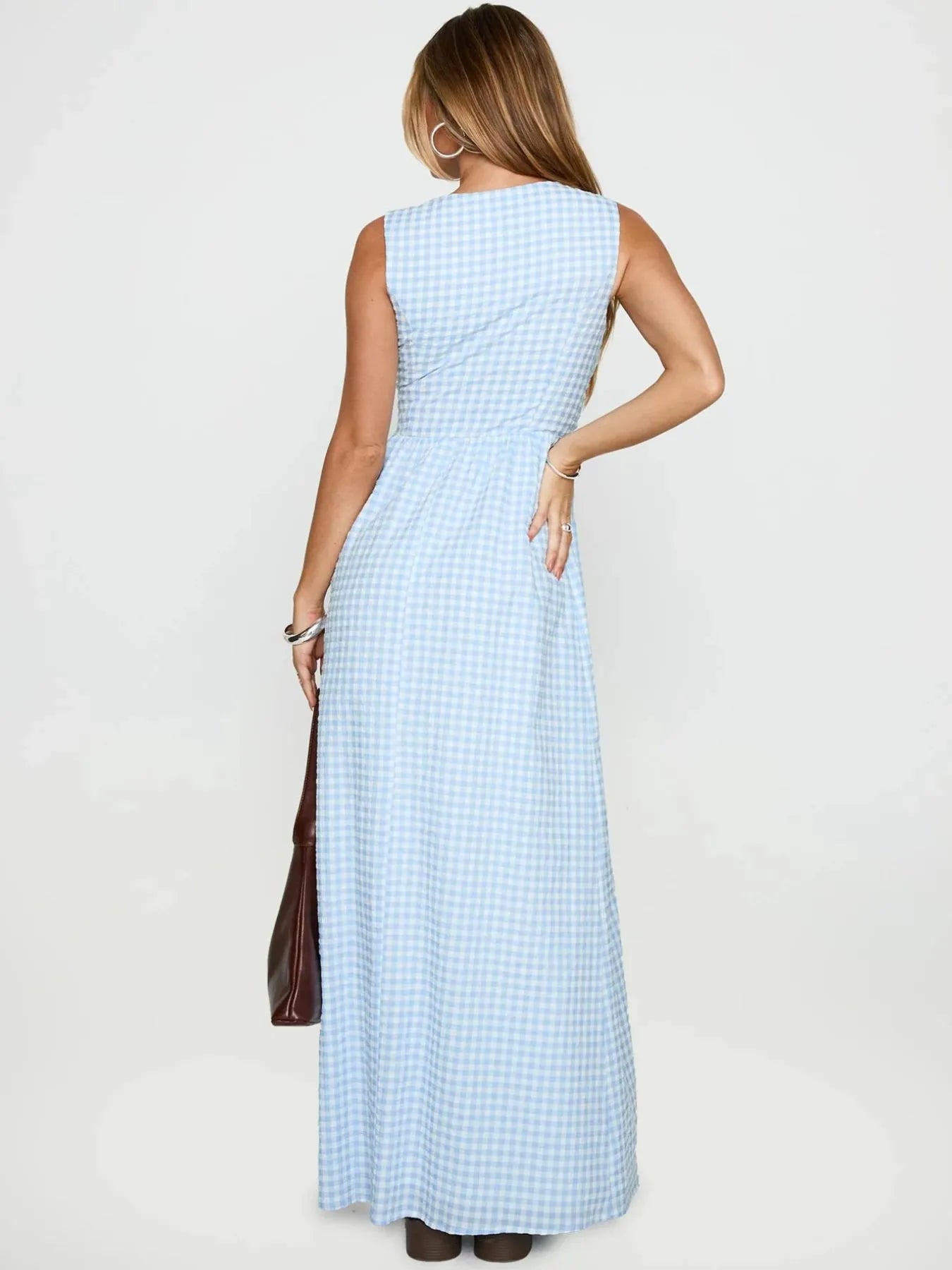 Back view of model wearing light blue gingham plaid tie front maxi dress sleeveless with brown tote bag